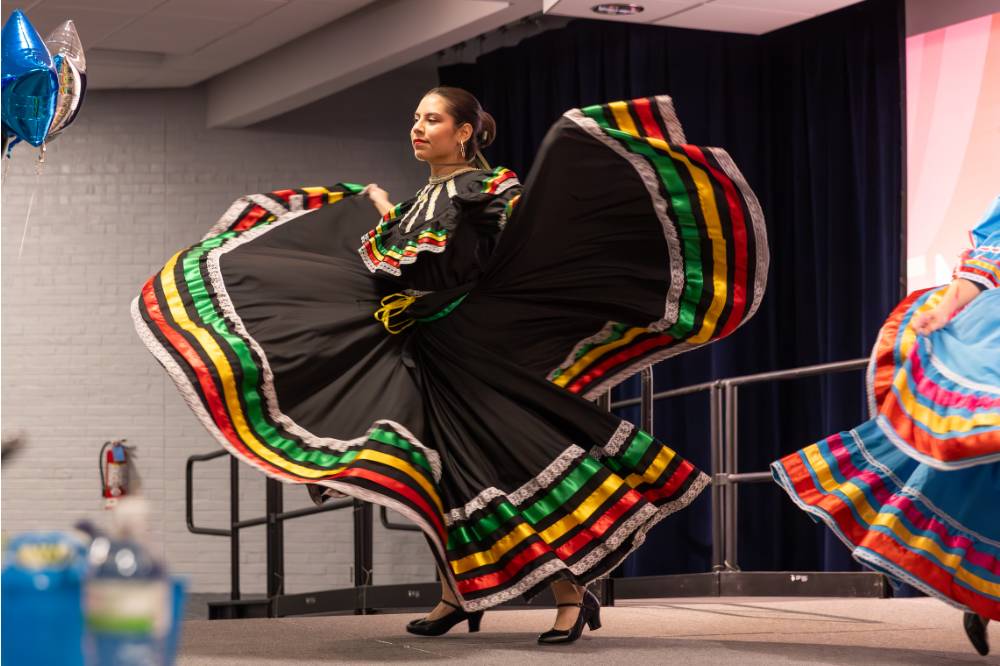 Monarcas ballet folklorico performer in black multicolored dress holding sides of skirt out on either side while dancing.